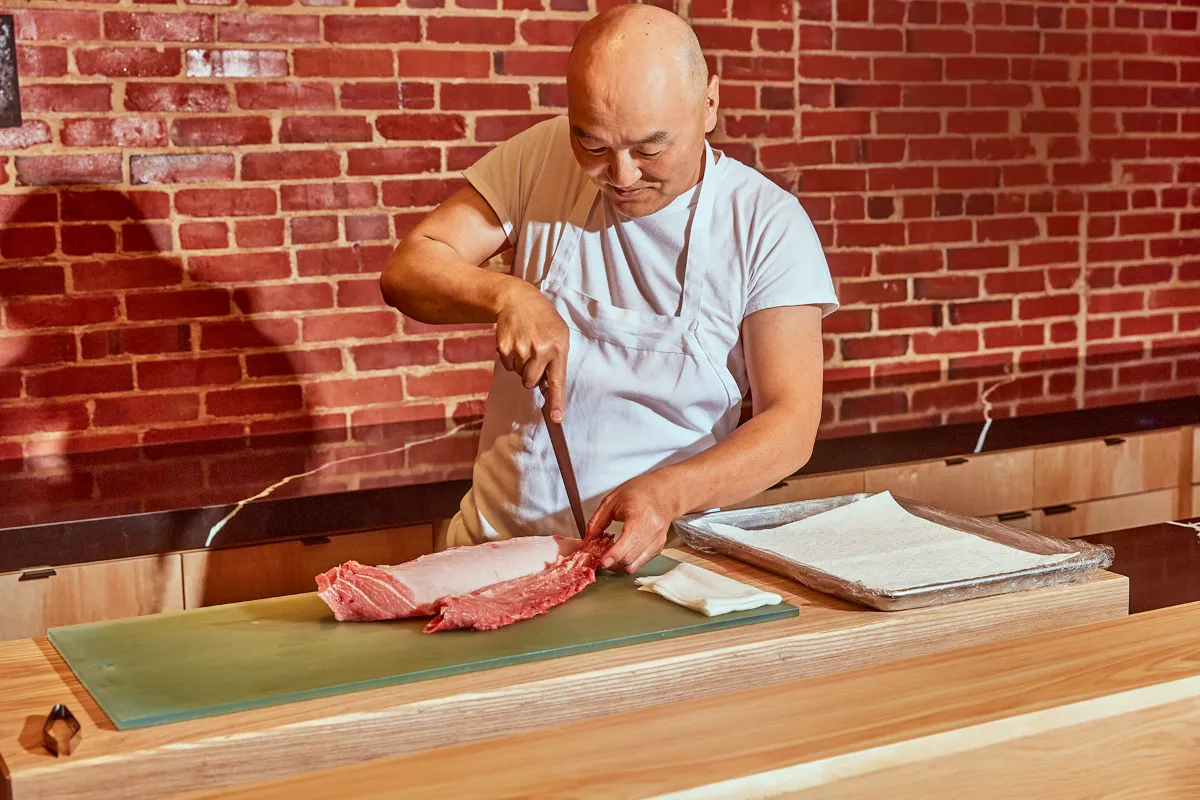 Chef do Tatsu cortando atum no balcão de omakase, com parede de tijolos ao fundo
