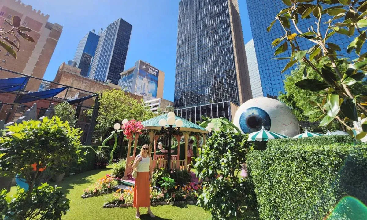 Turista em um jardim de Downtown Dallas, com o olho gigante e os prédios ao fundo