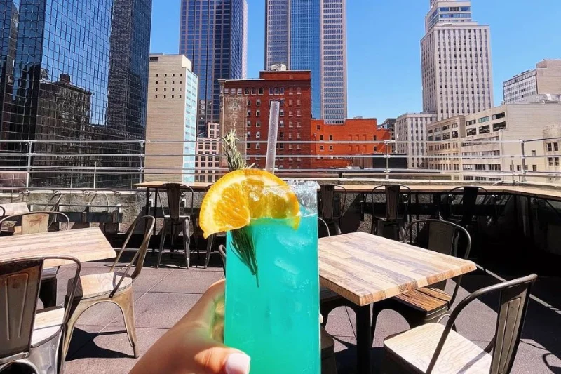 Drinque azul com fatia de laranja no terraço do Sky Blossom, com os arranha-céus do Downtown de Dallas ao fundo