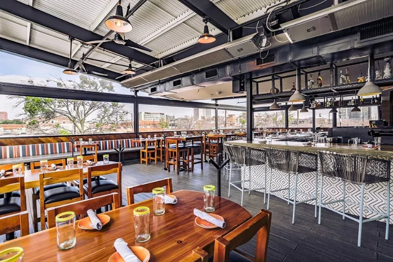Terraço do Vidorra no Deep Ellum, com mesas de madeira, bar com banquetas e vista aberta do bairro